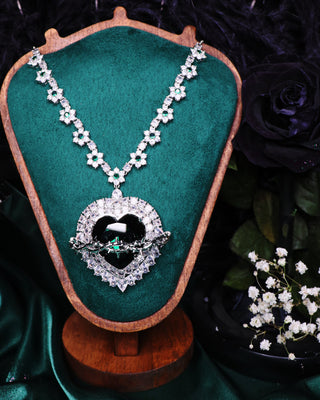 Elegant necklace with a heart-shaped pendant on a green velvet stand, surrounded by dark foliage and white flowers.
